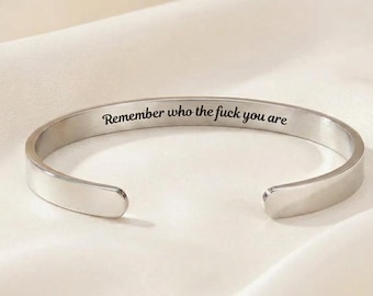 May include: A silver-toned metal cuff bracelet with an open design. The interior surface of the bracelet features the engraved black text "Remember who the fuck you are" in a clean, legible, sans-serif font style.