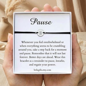 May include: A black cord bracelet with a circular silver charm featuring a pause symbol, presented in a white gift box. The card displays the text "Pause" and a message about taking a moment to breathe, ending with the URL brilagifts.etsy.com.