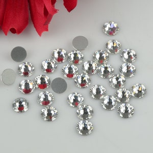 May include: A collection of clear crystal rhinestones of various sizes, scattered on a white surface. The rhinestones have a faceted design and sparkle in the light.