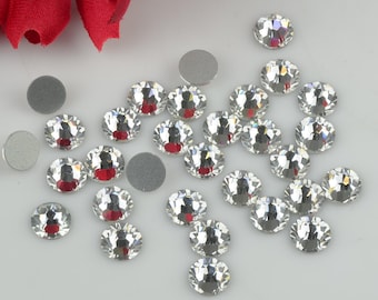 May include: A collection of clear crystal rhinestones of various sizes, scattered on a white surface. The rhinestones have a faceted design and sparkle in the light.