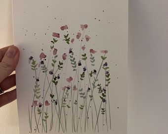 May include: Watercolor painting of wildflowers in shades of pink, purple, and green. The artwork features delicate stems and leaves, with a few dark speckles scattered across the white paper. The painting is held by a hand.