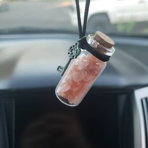 May include: A small glass bottle filled with pink Himalayan salt crystals hangs from a black string with a key charm. The bottle is hanging from the rearview mirror of a car.
