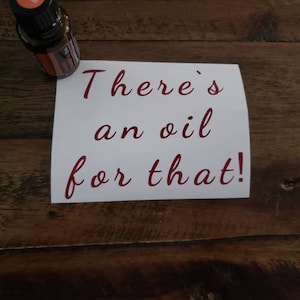 以下が含まれることがあります： 赤い筆記体で「There's an oil for that!」と書かれた白いカード。赤いキャップの小さな琥珀色のガラス瓶が木製の表面の上に置かれている。