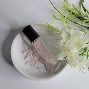 May include: A clear glass roller bottle with a black cap filled with pink crystals. The bottle is sitting on a white ceramic dish with more pink crystals scattered around it.