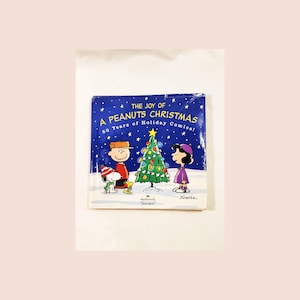 May include: A blue book cover with the title "The Joy of A Peanuts Christmas 50 Years of Holiday Comics!"  The cover features Charlie Brown, Snoopy, and Lucy standing in front of a decorated Christmas tree.