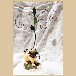 May include: A decorative figurine featuring a pug dog standing on a snow-covered base with a snowflake design. Above the pug is a black wire design with green leaves and red berries, resembling holly. The figurine is set against a white background.