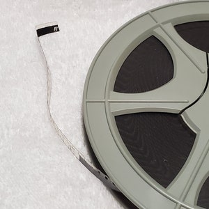 May include: A gray film reel with a black film strip extending from it. The film strip has the number "5" printed on a black and white label. The reel is on a white, textured surface.