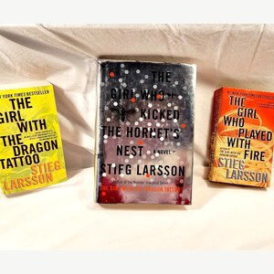 Pode incluir: São exibidos três livros de Stieg Larsson. As capas são amarelas, prateadas e laranja, com títulos como "The Girl with the Dragon Tattoo", "The Girl Who Kicked the Hornet's Nest" e "The Girl Who Played with Fire."