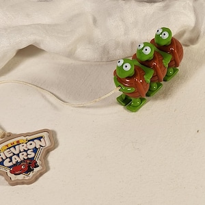 May include: A pull toy featuring three green turtles with brown shells connected in a line, attached by a white string to a tan-colored plastic tag with the text The Chevron Cars and an illustration of a red car printed on the front surface.
