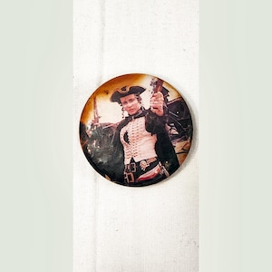 May include: A round button with a picture of a man in a white and gold uniform, wearing a black hat and holding a gun. The man is standing in front of a dark background.