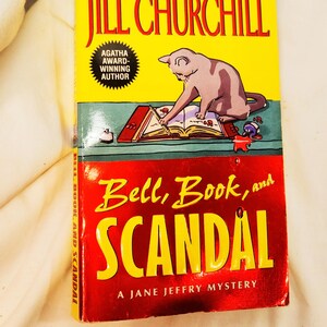 Pode incluir: Um livro de capa mole vermelho e amarelo intitulado "Bell, Book, and Scandal" de Jill Churchill. A capa do livro apresenta um gato sentado a uma mesa com um livro aberto e uma caneta na pata. O livro é um mistério de Jane Jeffry e a autora é uma autora vencedora do prêmio Agatha.