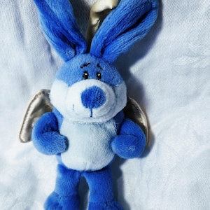 May include: A plush blue bunny with large ears, a white belly, and a blue nose. The stuffed animal has small black eyes and silver wings. The bunny is standing on a white surface.