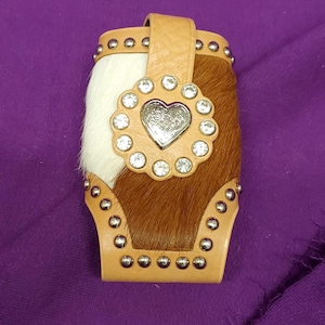 May include: A tan leather pouch featuring white and brown cowhide fur panels. The front is decorated with silver-tone metal studs along the edges and a central circular ornament containing a silver heart surrounded by clear, sparkling rhinestones.