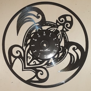 Può includere: Un orologio in vinile nero con un design a forma di tartaruga. Il quadrante dell'orologio è un disco in vinile nero più piccolo con numeri bianchi e un centro nero. Il testo "F.B. Creations" è stampato sul quadrante dell'orologio.