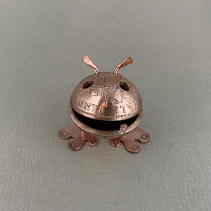 May include: A small copper frog sculpture made from a half penny coin. The frog has a wide mouth and two small antennae.