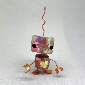 May include: A handcrafted metal robot sculpture with a square head and cylindrical body. The head is painted with a mix of pink, purple, and gold hues, featuring two black eye sockets. A gold heart is on the body. The robot has arms and feet.