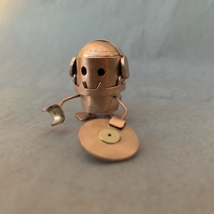 May include: A small, copper-coloured robot figurine with a round head, two arms, and two legs. The robot is wearing headphones and holding a small, round, copper-coloured record player.