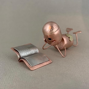 May include: A small, copper-coloured figurine of a person reading a book. The figurine is made of metal and has a detailed design. The book is open and has a silver-coloured interior.
