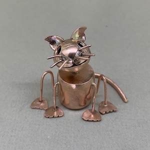 May include: A copper-coloured metal cat sculpture. The cat has a round body, four legs, a tail, and a head with ears and whiskers. The sculpture is sitting on a light green surface.