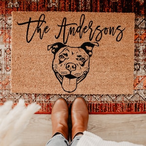 Peut inclure: Un paillasson rectangulaire en fibre de coco avec le texte "the Andersons" en cursive au-dessus d'un dessin au trait noir d'un visage de chien. Le paillasson est sur un tapis à motifs, et les pieds d'une personne en bottes marron sont visibles.