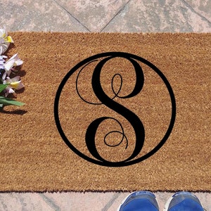 May include: A rectangular doormat made of natural coir fibre, featuring a large black letter "S" inside a circular frame. The mat is placed on a stone surface, with a bouquet of flowers to the left.