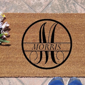 May include: A rectangular brown coir doormat with a black circular monogram design. The monogram features the letter "M" and the name "MORRIS" in a serif font. A bouquet of flowers is to the left.