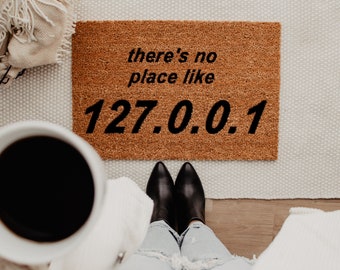 May include: A brown coir doormat with the text "there's no place like 127.0.0.1" in black. The image includes black boots, a white mug, and a beige blanket. The mat is rectangular and suitable for a doorway.
