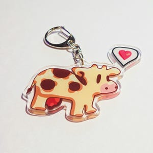 May include: A clear acrylic keychain featuring a cartoon cow with brown spots and a red udder. The cow has a speech bubble with a pink heart. The keychain has a silver clasp.