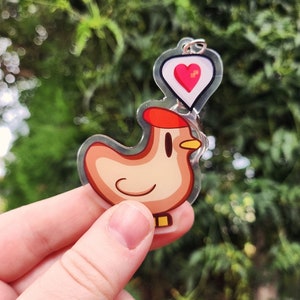May include: A clear acrylic keychain featuring a cartoon chicken with a red comb and a heart-shaped bubble. The chicken is light brown with a yellow beak and a smiling face. The heart has a pink heart inside. The keychain is held against a blurred green background.
