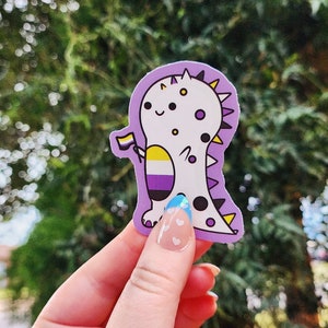 Puede incluir: Una pegatina de dinosaurio morado, blanco y amarillo con una pequeña bandera del orgullo. El dinosaurio sostiene la bandera en su mano derecha. El dinosaurio tiene una cola con manchas azules y blancas y un cuerpo blanco con manchas negras.