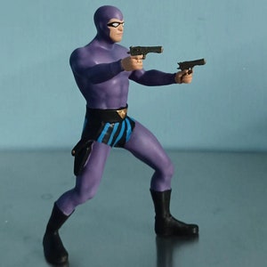 May include: A purple action figure of a superhero, wearing a mask and holding two black pistols. The figure has a black belt with blue stripes and black boots. The figure is in a dynamic pose, as if in action.