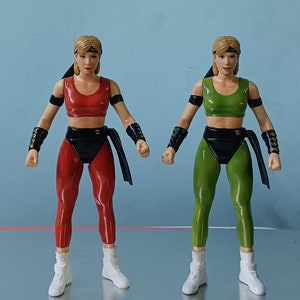 May include: Two action figures of a female character wearing different coloured outfits. The figure on the left is wearing a red outfit with black accents, while the figure on the right is wearing a green outfit with black accents. Both figures have blonde hair and are wearing white shoes.