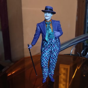 Pode incluir: Uma figura de ação do Joker em roxo e azul com um chapéu roxo, uma gravata borboleta verde e um terno xadrez roxo e azul. A figura está segurando uma bengala preta e está em pé em uma superfície dourada.