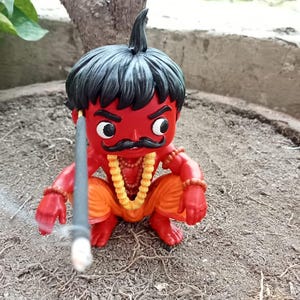 May include: A red-skinned figurine with black hair and a moustache, wearing orange trousers and yellow beaded necklaces. The figure is in a crouching position and holds a thin, dark grey incense stick that emits a small wisp of white smoke.