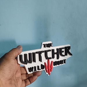 May include: A white 3D printed sign with the words "The Witcher Wild Hunt" in black lettering and a red Witcher logo.
