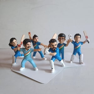 Op de afbeelding: Een verzameling miniatuur cricketspeler beeldjes. De beeldjes dragen blauwe uniformen met witte accenten en zijn in verschillende actieposities afgebeeld, met batten en ballen. De beeldjes staan op een wit oppervlak.