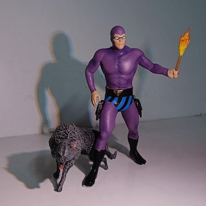 Könnte beinhalten: Lilafarbene Actionfigur eines Superhelden mit schwarzer Maske und einem blau-schwarz gestreiften Gürtel. Die Figur hält in einer Hand eine brennende Fackel und hat einen schwarzen Wolf zu seinen Füßen.