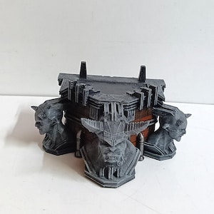 May include: A black and grey 3D printed box with three sculpted heads of horned creatures on the sides. The box is made of a textured material and has a dark grey finish.