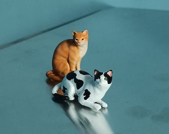May include: Two ceramic cat figurines. One is orange and white, sitting upright. The other is white with black spots, lying down. Both have detailed features and are set against a reflective surface with a blue background.