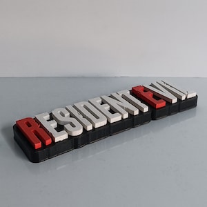 May include: A black and white 3D printed sign that reads "RESIDENT EVIL" with the "E" and "I" in red.