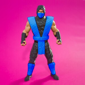 May include: A blue and black action figure of a ninja wearing a blue mask and a blue sash. The figure is standing on a pink background.
