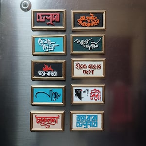 Pode incluir: Uma coleção de ímanes retangulares com molduras douradas. Cada íman apresenta texto colorido numa escrita única. As cores do texto incluem vermelho, branco, azul e laranja. Os ímanes são exibidos numa superfície metálica.