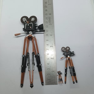 Puede incluir: Miniaturas de cámaras de cine vintage sobre trípodes. La cámara más grande mide aproximadamente 15 cm de alto, con una combinación de colores marrón y negro. Los trípodes tienen patas marrones y soportes negros. Una regla se coloca junto a las cámaras para la escala.