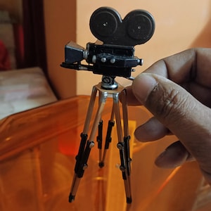 Puede incluir: Una cámara de cine en miniatura de color negro con dos carretes redondos en un trípode de madera.