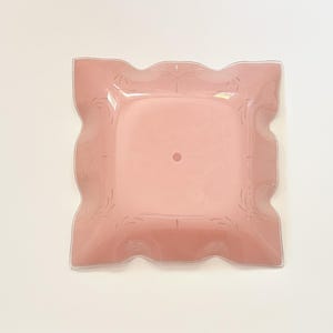 May include: A square, light pink glass plate with a scalloped edge and a central circular indentation. The plate features a subtle, etched floral pattern along its border, providing a delicate texture to the smooth, translucent pink surface.