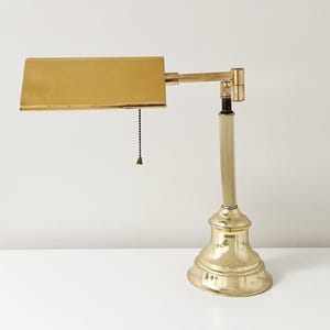 May include: A brass desk lamp with a rectangular shade and a pull chain. The lamp has a decorative base and an adjustable arm. The lamp is on a white surface.