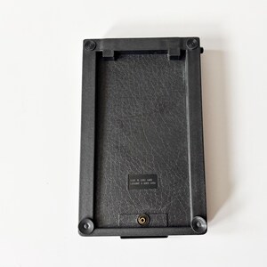Puede incluir: Un objeto rectangular negro con una superficie texturizada y esquinas redondeadas. El objeto tiene un peque&ntilde;o componente met&aacute;lico y cuatro pies circulares. El texto "MADE IN SRI LANKA" es visible en la superficie.