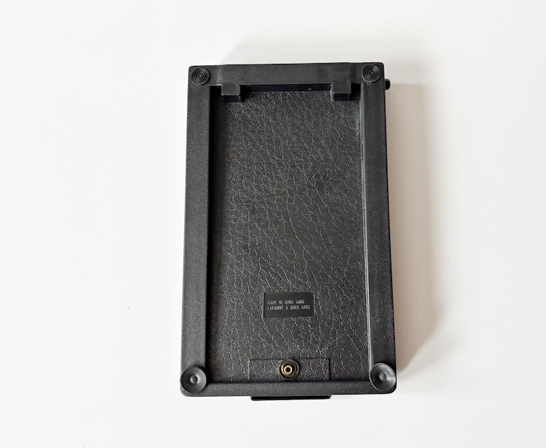 Puede incluir: Un objeto rectangular negro con una superficie texturizada y esquinas redondeadas. El objeto tiene un peque&ntilde;o componente met&aacute;lico y cuatro pies circulares. El texto "MADE IN SRI LANKA" es visible en la superficie.