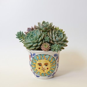 Può includere: Un vaso in ceramica bianca decorato con un sole giallo dipinto a mano con raggi blu, riempito con un gruppo di piante succulente verdi e viola chiaro. Il vaso presenta un bordo svasato e motivi floreali blu e rossi sui lati.