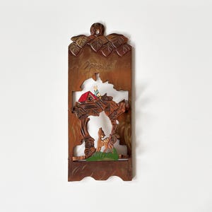 May include: A wooden wall hanging with a carved forest scene. The brown frame features a house with a red roof, a deer, and a bird. The word "Orest" is carved at the top. The piece is approximately 30 cm tall.